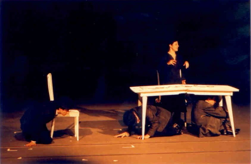 Il Processo — tavolo-tribunale, protagonista in piedi e tre figure inginocchiate per terra, scena minimale, scenografia Cristian Taraborrelli, regia Corsetti, TNSJ Porto PoNTI'99