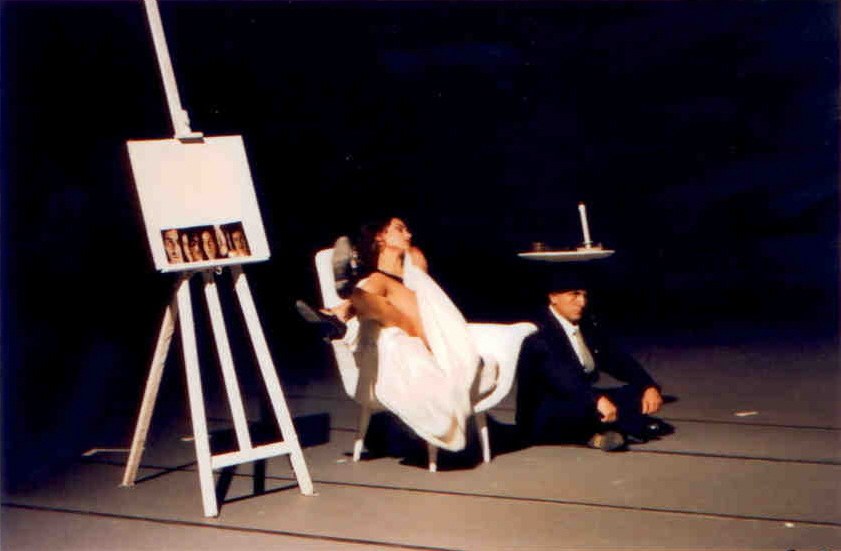 Il Processo — atelier di Titorelli, cavalletto con polaroid + donna in lenzuolo + uomo con candela su piatto, citazione magrittiana, scene Cristian Taraborrelli